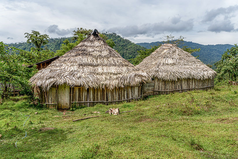 Zwei traditionelle Rundhütten mit markanten kegelförmigen Strohdächern und Wänden aus gespaltenen Holzpfählen stehen auf einer grünen Lichtung vor einer bewaldeten Bergkulisse. Puerto Viejo de Talamanca Bribri