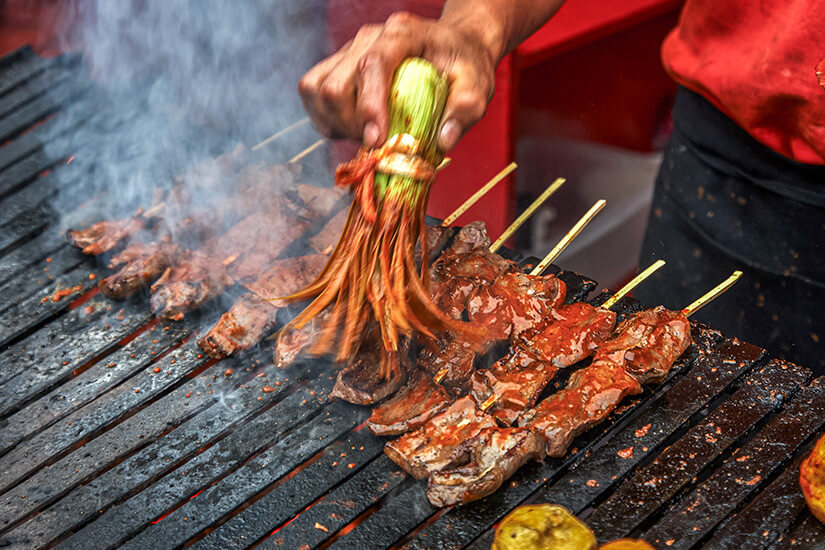 Eine Hand bestreicht auf einem rauchenden Holzkohlegrill aufgespießte Fleischstücke mit einer würzigen roten Marinade, die mithilfe eines Bündels frischer Frühlingszwiebeln als natürlichem Pinsel aufgetragen wird. Peruanisches Essen Streetfood
