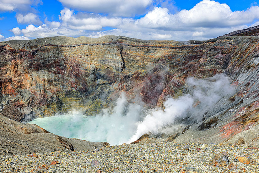 Ein Blick direkt in den aktiven Naka-Krater des Aso-Vulkans offenbart einen türkis schimmernden Kratersee, aus dem dichte Dampfwolken aufsteigen. Die farbenprächtigen Gesteinsschichten in Rot-, Gelb- und Grautönen zeigen die unglaubliche geologische Vielfalt dieses Ortes. Es ist ein surrealer, fast außerirdischer Anblick, der unter den Kumamoto Sehenswürdigkeiten seinesgleichen sucht. Kumamoto Japan Krater