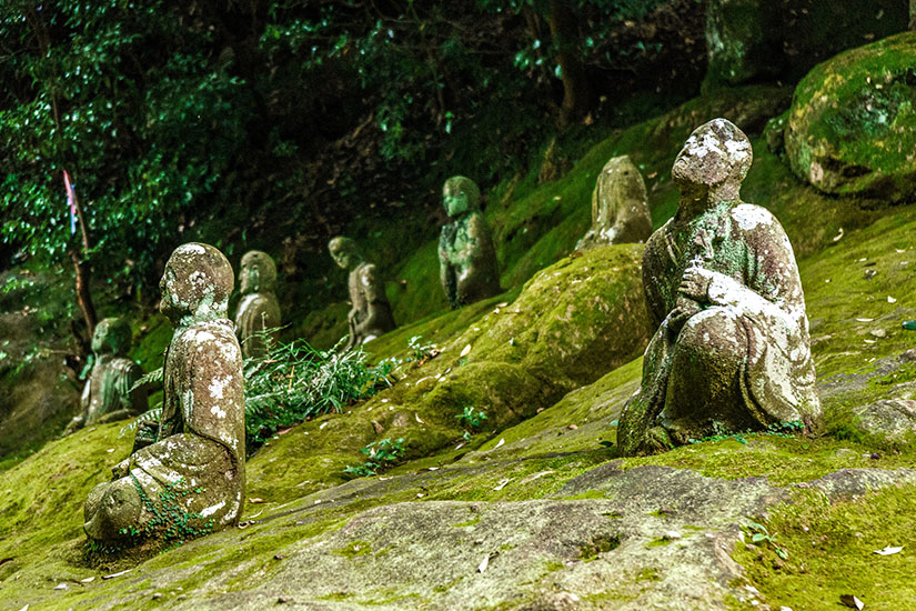 Eine Reihe verwitterter, moosbewachsener Steinstatuen sitzt friedlich auf einem grünen Felshang im mystischen Reigando-Höhlentempel. Die jahrhundertealten Figuren strahlen eine fast überirdische Ruhe aus und schaffen eine fast magische Atmosphäre zwischen Stein und Natur. Dieser spirituelle Ort gehört zu den geheimnisvollsten Kumamoto Sehenswürdigkeiten und war einst Rückzugsort des legendären Samurai Miyamoto Musashi. Kumamoto Japan Unganzenji