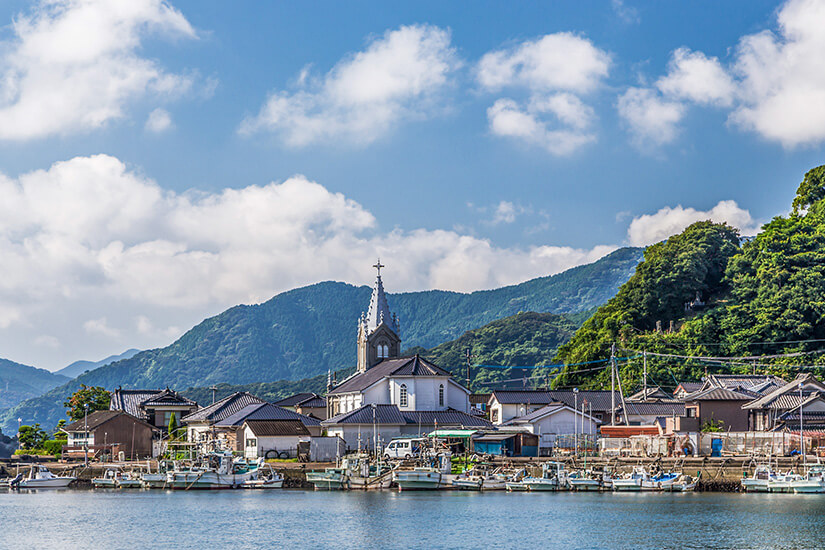 Die markante Sakitsu-Kirche mit ihrem spitzen Glockenturm ragt zwischen traditionellen japanischen Häusern aus einem malerischen Fischerdorf empor und spiegelt sich im ruhigen Hafenwasser. Dieser ungewöhnliche Anblick einer gotisch anmutenden Kirche zwischen Fischerbooten erzählt die bewegende Geschichte der versteckten Christen Japans. Die Region Amakusa gehört zum UNESCO-Weltkulturerbe und ist ein einzigartiges Highlight unter den Sehenswürdigkeiten von Kumamoto. Kumamoto Japan Kirche