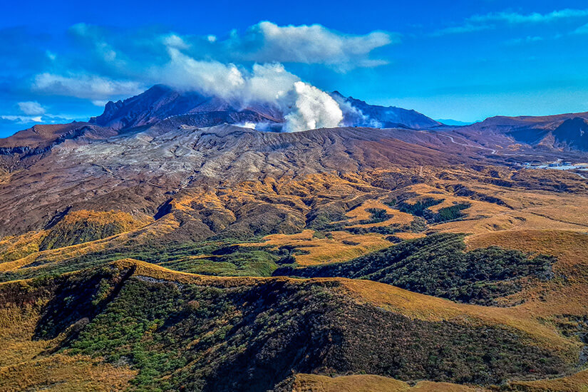 Der majestätische Aso-Vulkan thront über der weiten, herbstlich gefärbten Hügellandschaft und stößt eine sichtbare Rauchwolke in den strahlend blauen Himmel. Die wellenförmigen Hänge mit ihrem goldbraunen Gras und den grünen Vegetationsinseln zeigen die einzigartige Caldera-Landschaft, die Kumamoto in Japan weltberühmt gemacht hat. Dieser Anblick gehört zweifellos zu den eindrucksvollsten Naturschauspielen, die man in Kyushu erleben kann. Kumamoto Japan Aso