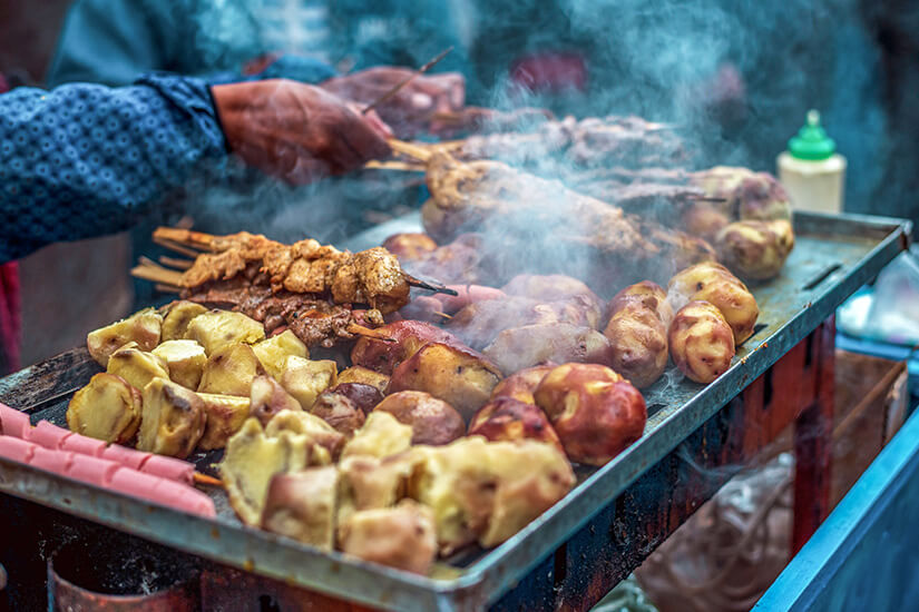 Ein dampfender Straßengrill ist beladen mit gegrillten Kartoffeln, Fleischspießen und weiteren herzhaften Snacks, während eine Verkäuferin die Speisen auf einem mobilen Grillwagen zubereitet. Peruanisches Essen Tradition