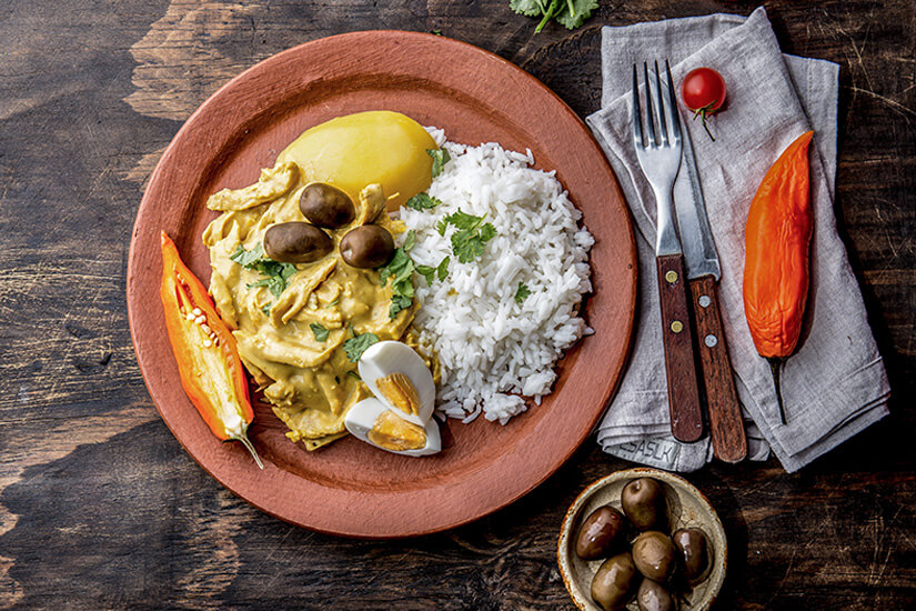 Auf einem rustikalen Tonteller ist das peruanische Nationalgericht angerichtet: zartes Hühnerfleisch in einer cremig-gelben Ají-Amarillo-Soße, garniert mit schwarzen Oliven, gekochtem Ei und frischem Koriander, begleitet von weißem Reis und einer ganzen Kartoffel. Peruanisches Essen Gallina