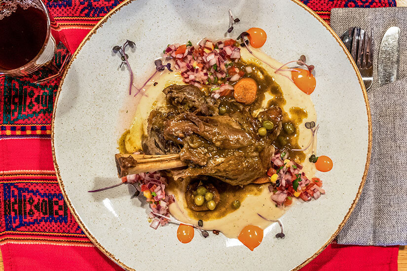 Die gehobene Präsentation auf dem handgetöpferten Teller, kombiniert mit dem farbenfrohen traditionellen Andentischtuch im Hintergrund, verrät ein gehobenes Restaurant in Peru, das die moderne Landesküche mit klassischen Zutaten neu interpretiert. Peruanisches Essen Lamm