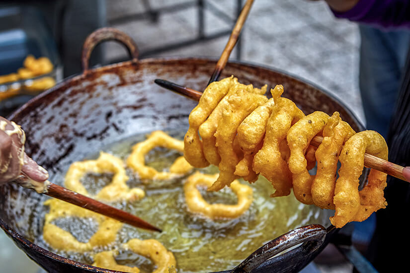 In einem großen, schwarz geschwärzten Wok werden goldbraune Teigringe in heißem Öl ausgebacken, während ein Stäbchen einen fertig gebratenen Ring aus dem sprudelnden Fett hebt. Peruanisches Essen Picarones