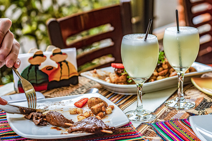Zwei cremig-weiße Pisco Sour Cocktails in eleganten Kelchgläsern dominieren den Tisch eines peruanischen Restaurants, auf dem gegrilltes Fleisch mit Andenkartoffeln auf einer bunten Tischdecke serviert wird. Peruanisches Essen Pisco Sour
