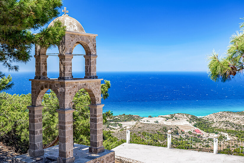 Ein steinerner Glockenturm mit offenen Bögen steht auf einer Anhöhe mit Blick auf das tiefblaue Meer. Pinien rahmen die Szene ein, während unten eine helle Küstenlandschaft zu erkennen ist. Kos oder Rhodos Einleitung