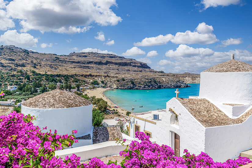 Weiße Häuser mit runden Dächern liegen oberhalb einer türkisfarbenen Bucht. Pinke Blumen im Vordergrund setzen starke Farbakzente vor der felsigen Küstenlandschaft. Rhodos oder Kos Standort