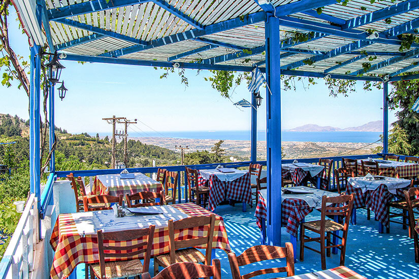 Eine offene Terrasse mit blau gestrichenen Holzbalken dient als Restaurant mit Blick aufs Meer. Die Tische sind mit karierten Tischdecken gedeckt, während im Hintergrund eine hügelige Küstenlandschaft zu sehen ist. Rhodos oder Kos Kueche