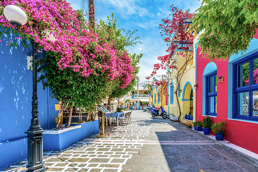 Eine schmale Gasse mit bunt gestrichenen Häusern wird von üppigen pinken Blüten und grünen Pflanzen geschmückt. Kleine Tische und Stühle stehen draußen und verleihen der Straße eine gemütliche Atmosphäre. Im Hintergrund sind ein Motorroller und weitere Gebäude unter blauem Himmel zu erkennen. Kos oder Rhodos Preise