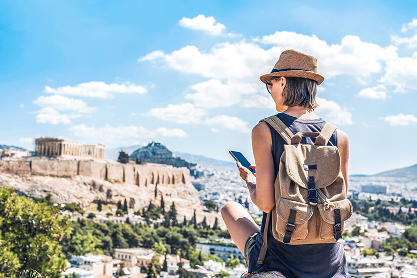 Eine junge Reisende mit Strohhut, Sonnenbrille und Rucksack sitzt auf einem Aussichtspunkt und betrachtet mit ihrem Smartphone das beeindruckende Panorama der Athener Akropolis mit dem Parthenon, das majestätisch über der weitläufigen Stadtlandschaft thront. Internet im Ausland eSim Frau