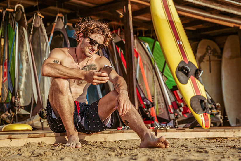 Ein entspannter junger Mann mit lockigem Haar, Sonnenbrille und Shorts sitzt lässig im Sand und scrollt auf seinem Smartphone, während hinter ihm eine bunte Sammlung von Surfboards, Segeln und Ausrüstung an einem Strandverleih lehnt. Internet im Ausland eSim Surfer