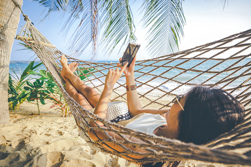 Eine junge Frau liegt entspannt in einer Hängematte zwischen Palmen am Strand und scrollt auf ihrem Smartphone, während das türkisblaue Meer im Hintergrund sanft in der Sonne glitzert. Internet im Ausland eSim Haengematte