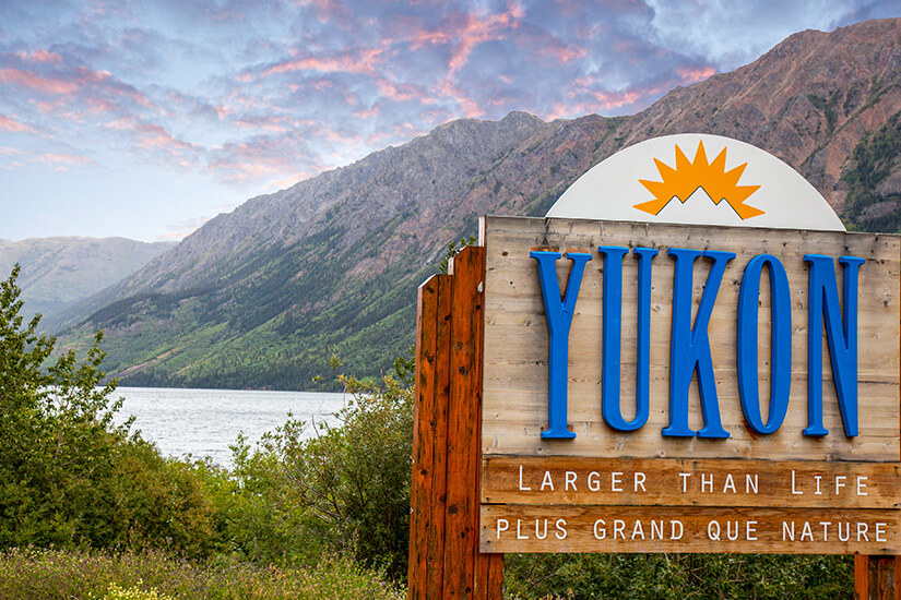 Ein großes Holzschild mit der Aufschrift „Yukon“ steht vor einer beeindruckenden Berglandschaft. Im Hintergrund sind ein See, bewaldete Hänge und hohe Berge unter einem leicht rosafarbenen Himmel zu sehen. Yukon Schild