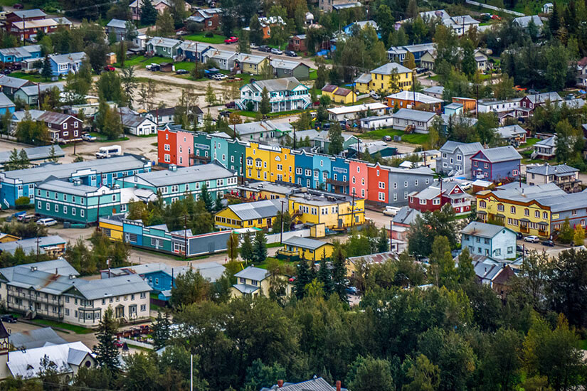 Eine Kleinstadt mit vielen bunten Häuserfassaden erstreckt sich zwischen dichten Bäumen und kleinen Straßen. Die Gebäude sind in kräftigen Farben wie Gelb, Blau, Rot und Grün gestrichen und verleihen dem Ort ein lebendiges Erscheinungsbild. Kanada Dawson City