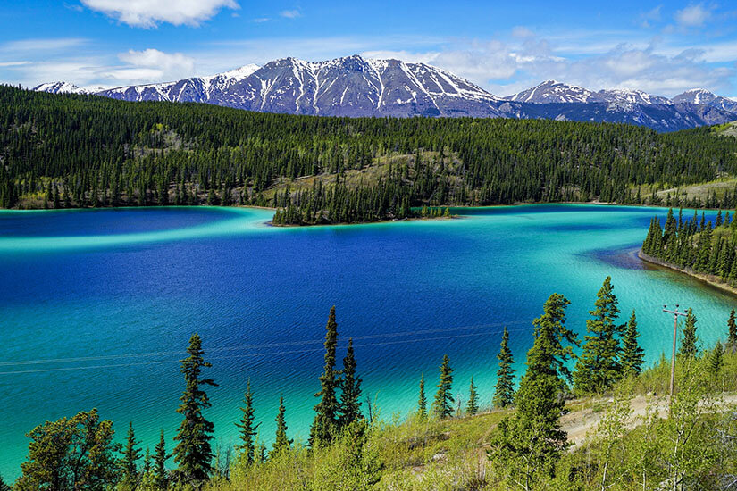 Ein leuchtend türkisfarbener See schlängelt sich durch eine Landschaft aus dichten Nadelwäldern und Bergen mit schneebedeckten Gipfeln. Im Vordergrund führt eine Straße entlang des Hangs oberhalb des Sees. Yukon Emerald Lake