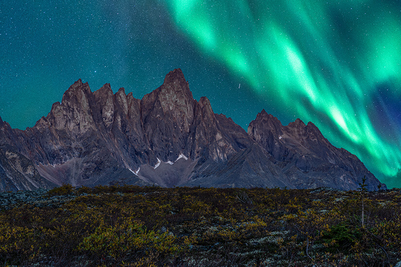 Über einer schroffen Bergkette tanzen leuchtend grüne Polarlichter am nächtlichen Himmel. Zwischen den Felsen und der dunklen Landschaft sind einzelne Sterne sichtbar. Yukon Polarlichter