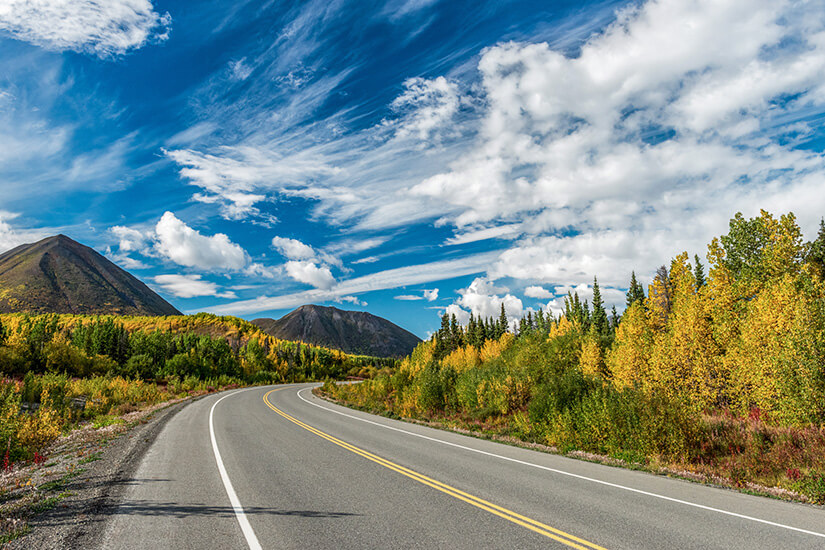 Eine lange Straße führt durch eine Landschaft mit herbstlich gefärbten Bäumen und bewaldeten Hügeln. Im Hintergrund ragen markante Berge unter einem Himmel voller verstreuter Wolken auf. Yukon Highway