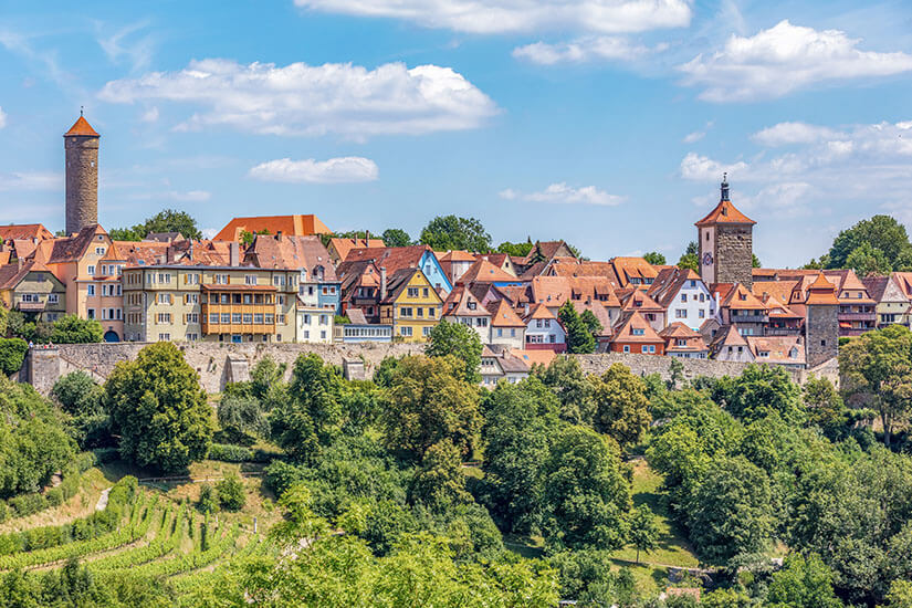 Die Altstadt von Rothenburg ob der Tauber mit bunten Fassaden und roten Dächern ist hinter der Stadtmauer zu sehen. Davor thronen grüne Bäume. Altstadt Deutschland Rothenburg