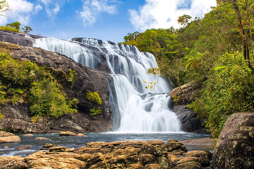 Ein prächtiger Wasserfall ergießt sich zwischen grüner Vegetation über dunkle Felsen in ein Wasserbecken. Nationalpark Sri Lanka Horton
