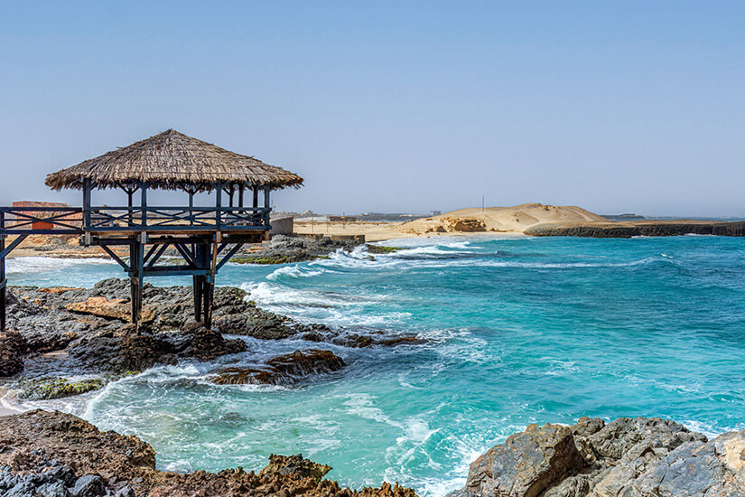 Türkisblaues Meer mit weißer Gischt schlägt gegen eine dunkle, felsige Küste, an der eine hölzerne Aussichtsplattform mit Strohdach auf Stelzen steht, während im Hintergrund eine karge Wüstenlandschaft unter hellem Himmel zu sehen ist. Boa Vista oder Sal Meer und Klippen