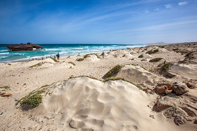 Das rostige Wrack des Schiffes Cabo Santa Maria liegt im türkisblauen Wasser direkt am Ufer eines weitläufigen Sandstrandes mit hellen Dünen und vereinzelten grünen Pflanzen unter einem leicht bewölkten blauen Himmel. Boa Vista oder Sal Strand mit Wrack