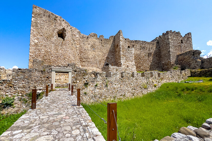 Eine alte Festungsruine aus hellem Stein erhebt sich unter einem wolkenlosen blauen Himmel. Ein schmaler gepflasterter Weg führt durch eine grüne Wiese direkt zu den massiven Mauern. Patras Kastell