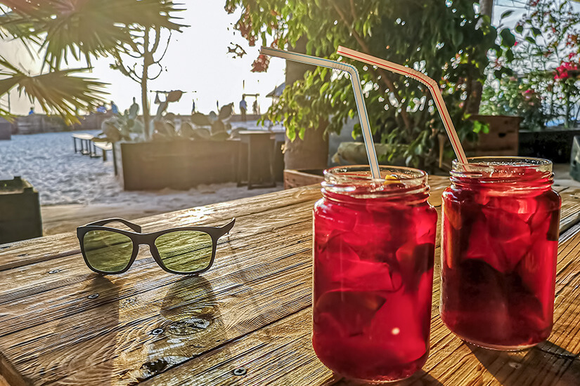 Zwei Gläser mit einem tiefroten Kaltgetränk, Eiswürfeln und Strohhalmen stehen neben einer Sonnenbrille auf einem rustikalen Holztisch an einem hellen Sandstrand bei tiefstehender Sonne. Boa Vista oder Sal Cocktails am Strand
