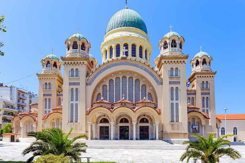 Eine große Kathedrale mit mehreren Kuppeln und hohen Türmen steht auf einem weiten Platz. Die helle Fassade ist reich verziert und verbindet klassische sowie byzantinisch wirkende Architekturelemente. Vor dem Gebäude wachsen Palmen unter einem strahlend blauen Himmel. Patras Kathedrale
