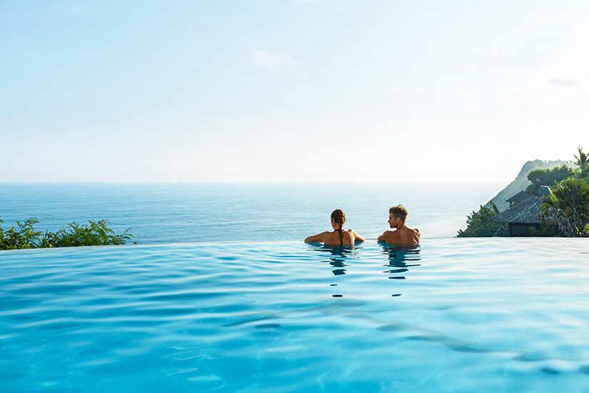 Ein Pärchen genießt innige Zweisamkeit in einem Infinity-Pool, dessen Wasserlinie nahtlos mit dem weiten, dunstigen Ozean am Horizont zu verschmelzen scheint, flankiert von üppigen Klippen mit tropischer Vegetation. Packliste Wellnessurlaub Paar