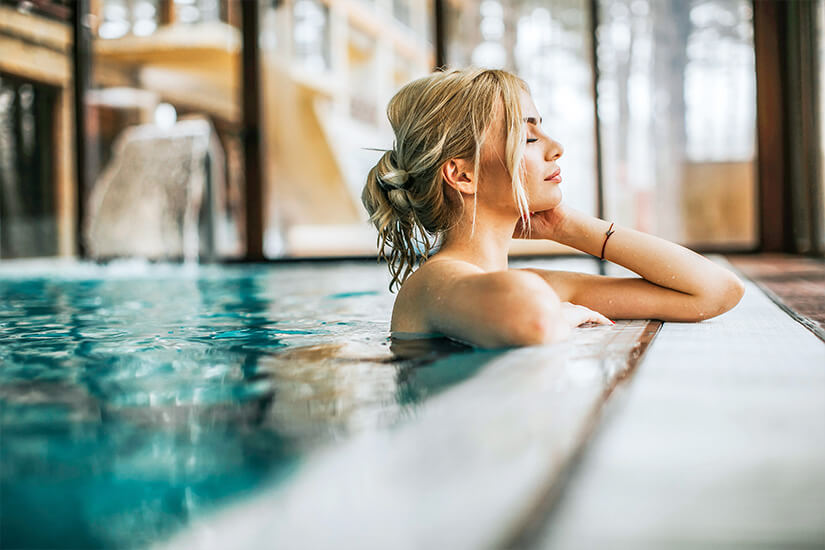Die Augen geschlossen, lehnt eine junge Frau mit hochgestecktem Haar entspannt am Rand eines dampfenden Innenpools und genießt in völliger Stille die wohlige Wärme des Thermalwassers. Packliste Wellnessurlaub Pool