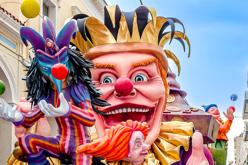 Bunte und überdimensionale Karnevalsfiguren mit clownartigen Gesichtern dominieren die Straße. Auffällige Farben, groteske Masken und fantasievolle Kostüme sorgen für eine ausgelassene und verspielte Stimmung. Im Hintergrund sind weitere Figuren und Gebäude einer Stadt zu erkennen. Patras Karneval