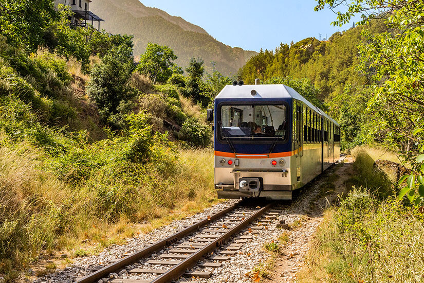 Ein kleiner Zug fährt auf schmalen Gleisen durch eine dicht bewachsene Berglandschaft. Hohe Bäume, trockenes Gras und grüne Hügel umgeben die Strecke auf beiden Seiten. Peloponnes Bahn