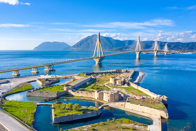Eine lange Schrägseilbrücke spannt sich über ein tiefblaues Meer zwischen Küsten und Bergen. Im Vordergrund liegen historische Festungsanlagen und Wassergräben auf einer kleinen Landzunge. Griechenland Bruecke