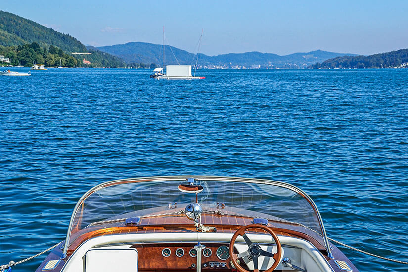 Ein Boot fährt über den blau leuchtenden Wörthersee und blickt dabei auf andere Boote und Ortschaften an fernen Ufern. Velden Oesterreich See