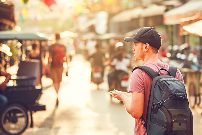 Mit Rucksack, Cap und Smartphone in der Hand steht ein junger Reisender auf einer belebten, von warmem Gegenlicht durchfluteten Straße und orientiert sich inmitten des pulsierenden Treibens einer asiatischen Metropole. Essen Kambodscha Tourist