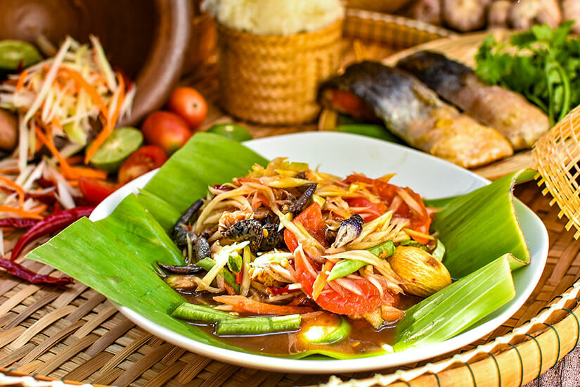 Auf einem Bananenblatt angerichtet präsentiert sich ein bunter Papayasalat mit Tomaten, langen Bohnen, getrockneten Krebsen und würziger Sauce, umgeben von typischen Beilagen wie Klebreis im Bambuskorb und gegrilltem Fisch. Essen Kambodscha Bok