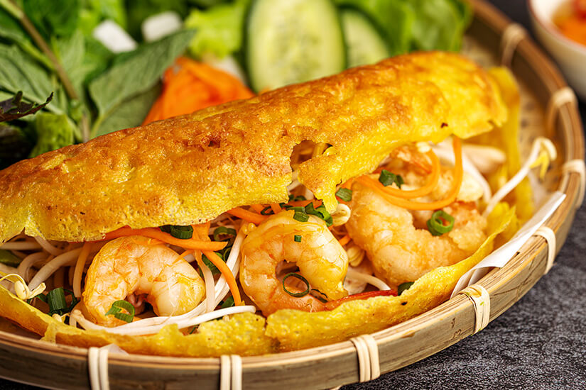 Knusprig goldbraune Reismehlcrêpes gefüllt mit saftigen Garnelen, Sojasprossen, Karotten und Frühlingszwiebeln sind in einem geflochtenen Bambuskorb angerichtet, begleitet von frischem Salatblättern, Gurke und Minze. Essen Kambodscha Banh Chev