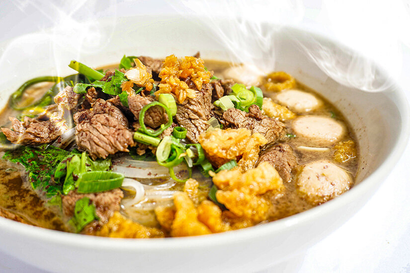 Dampfend heiß präsentiert sich eine aromatische Suppe mit zarten Fleischscheiben, knusprig frittierten Einlagen, Frühlingszwiebeln und frischem Koriander in einem dunklen, würzigen Fond. Essen Kambodscha Kuy Teav