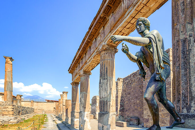 Zwischen antiken Säulen und Ruinen steht eine bronzefarbene Statue einer menschlichen Figur mit ausgestreckten Armen. Im Hintergrund sind Berge unter einem klaren blauen Himmel zu erkennen. Antike Stadt Pompeji