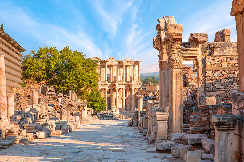 Eine breite antike Straße führt durch beeindruckende Ruinen aus Stein und Marmor. Am Ende der Straße steht ein prachtvolles Bauwerk mit mehreren Säulen und kunstvollen Details. Tuerkei Ephesos