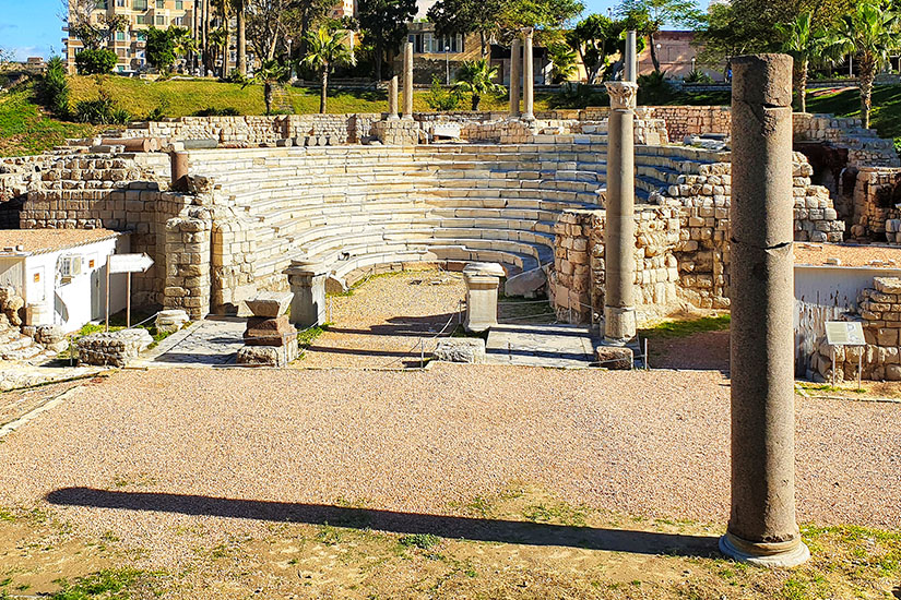 Ein antikes Amphitheater mit halbkreisförmigen Sitzreihen liegt zwischen alten Steinmauern und Säulenresten. Im Hintergrund stehen moderne Gebäude und Palmen, die einen Kontrast zur antiken Architektur bilden. Antike Staedte Alexandria
