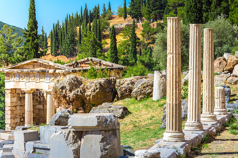 Zwischen antiken Ruinen stehen mehrere hohe Marmorsäulen vor einer hügeligen Landschaft mit Zypressen und Olivenbäumen. Griechenland Delphi
