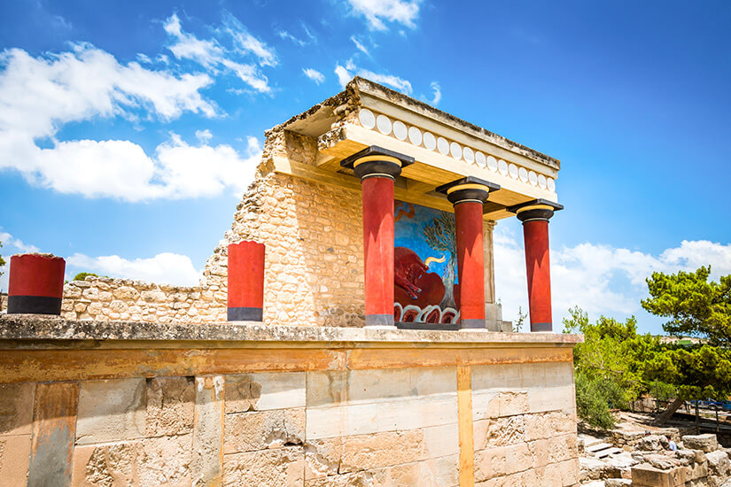 Ein antikes Bauwerk mit auffälligen roten Säulen steht unter einem strahlend blauen Himmel. Zwischen den Säulen ist ein farbenfrohes Wandbild zu sehen, das an minoische Kunst erinnert. Kreta Knossos
