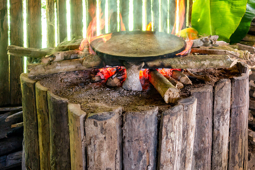 Auf einem rustikalen, aus rohen Holzstämmen und Lehm gefertigten Feuerherd lodern Flammen unter einer flachen Tonplatte, während im Hintergrund Bambuspfähle und ein Bananenblatt eine tropische Umgebung andeuten. Ecuador Essen Ofen
