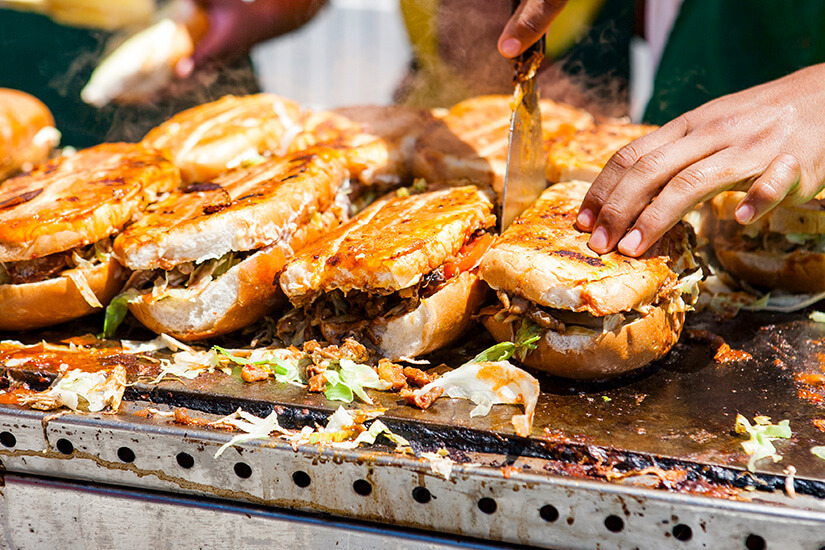 Auf einer heißen Straßengrill-Platte werden mehrere großzügig belegte, goldbraun geröstete Sandwiches mit Fleisch, Salat und Soßen von flinken Händen zubereitet und aufgeschnitten. Ecuador Essen Streetfood