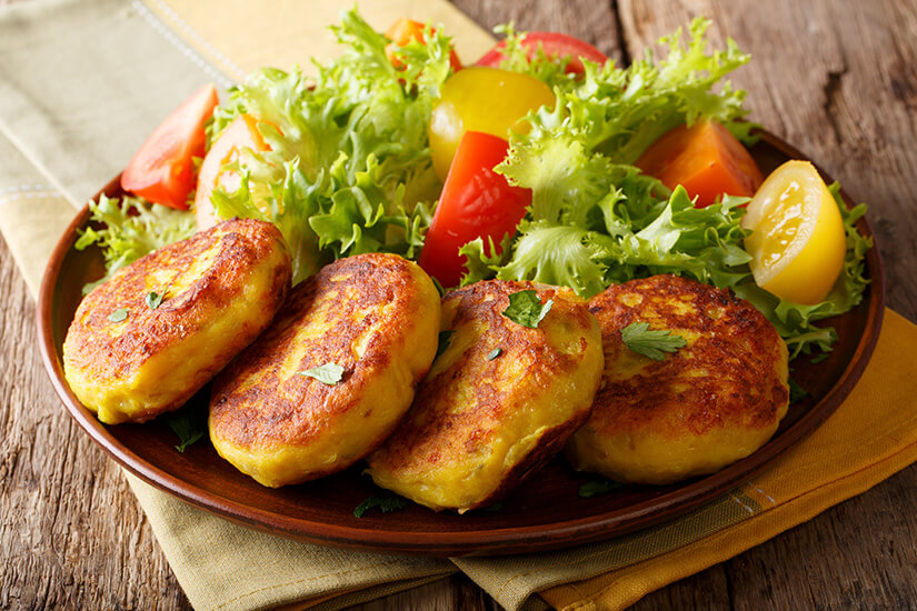 Goldbraun gebratene Kartoffelfrikadellen mit knuspriger Außenhülle und weich-cremigem Inneren sind auf einem rustikalen Tonteller angerichtet, begleitet von einem frischen Blattsalat mit roten und gelben Tomaten. Ecuador Essen Llapingachos