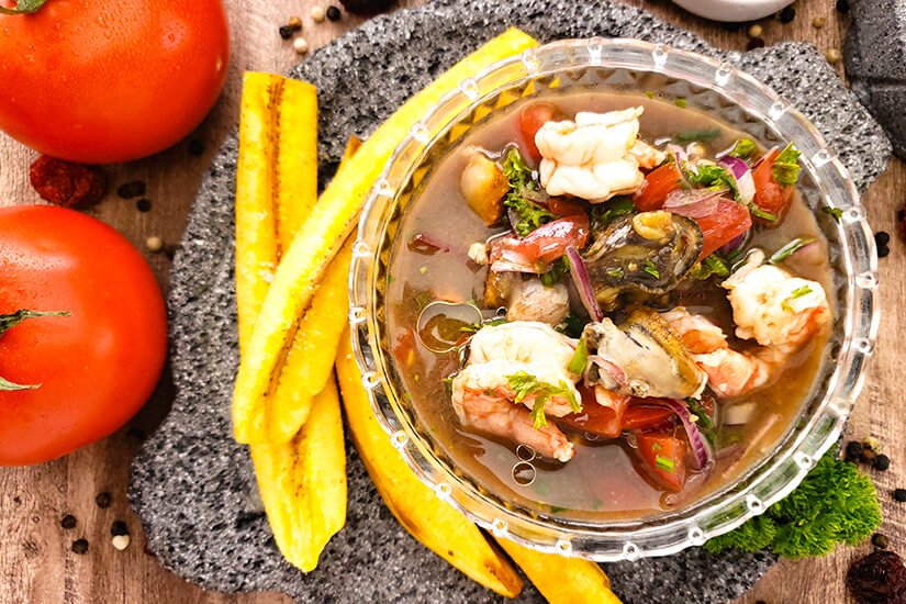 In einer gläsernen Schale schwimmen Garnelen, Muscheln, Tomaten und rote Zwiebeln in einem würzigen Meeresfrüchte-Ceviche, begleitet von gebratenen Kochbananenscheiben als typische Beilage. Ecuador Essen Ceviche