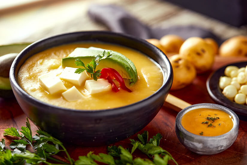 In einer dunklen Keramikschale dampft eine sattgelbe, cremige Suppe, garniert mit Avocado, weißen Käsewürfeln, roter Chili und frischem Koriander, begleitet von Andenkartoffeln, weißem Mais und einer kleinen Schale Ají. Ecuador Essen Kartoffelsuppe